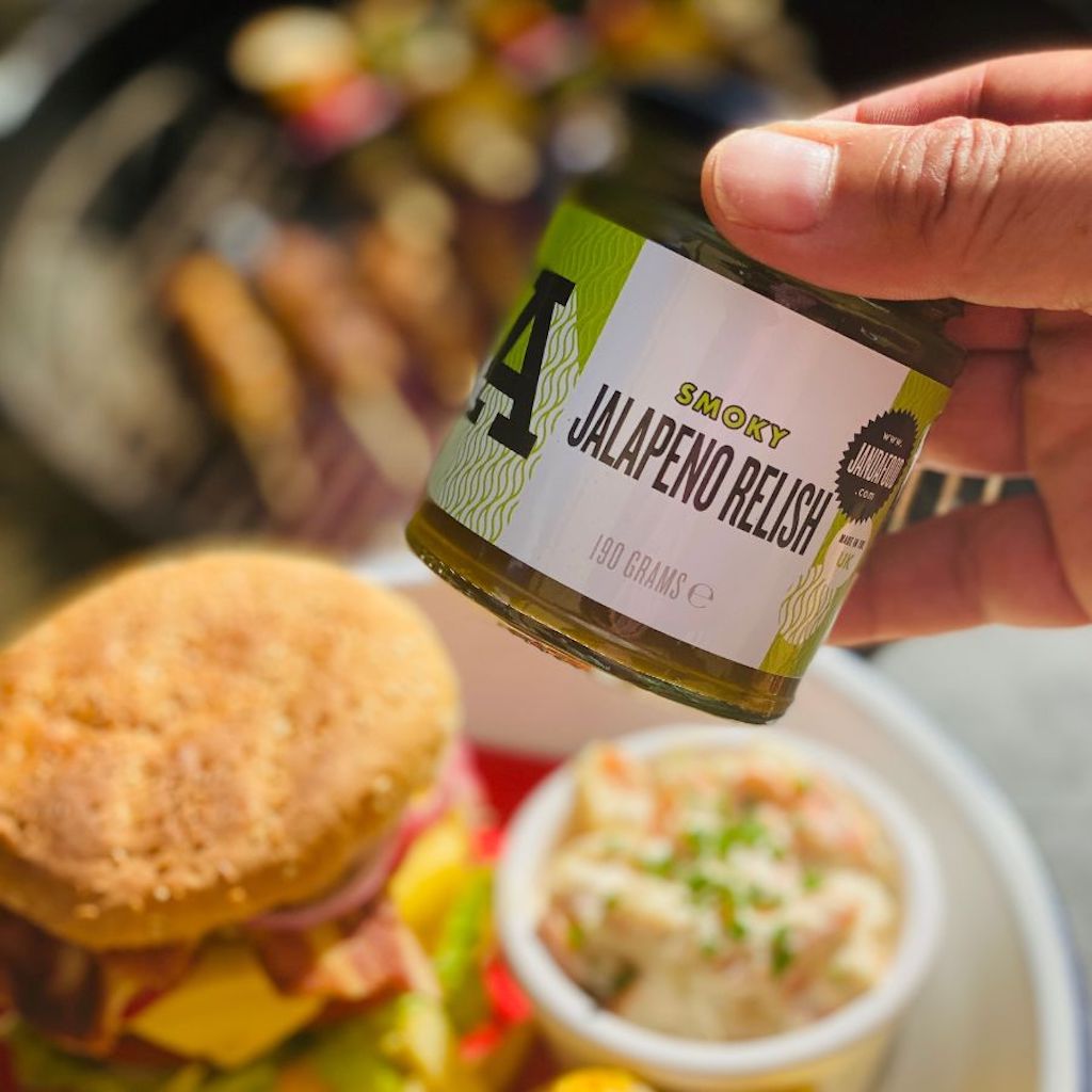 Hand holding a jar of 'Smoky Jalapeno Relish' with a blurred background of a burger and salad.