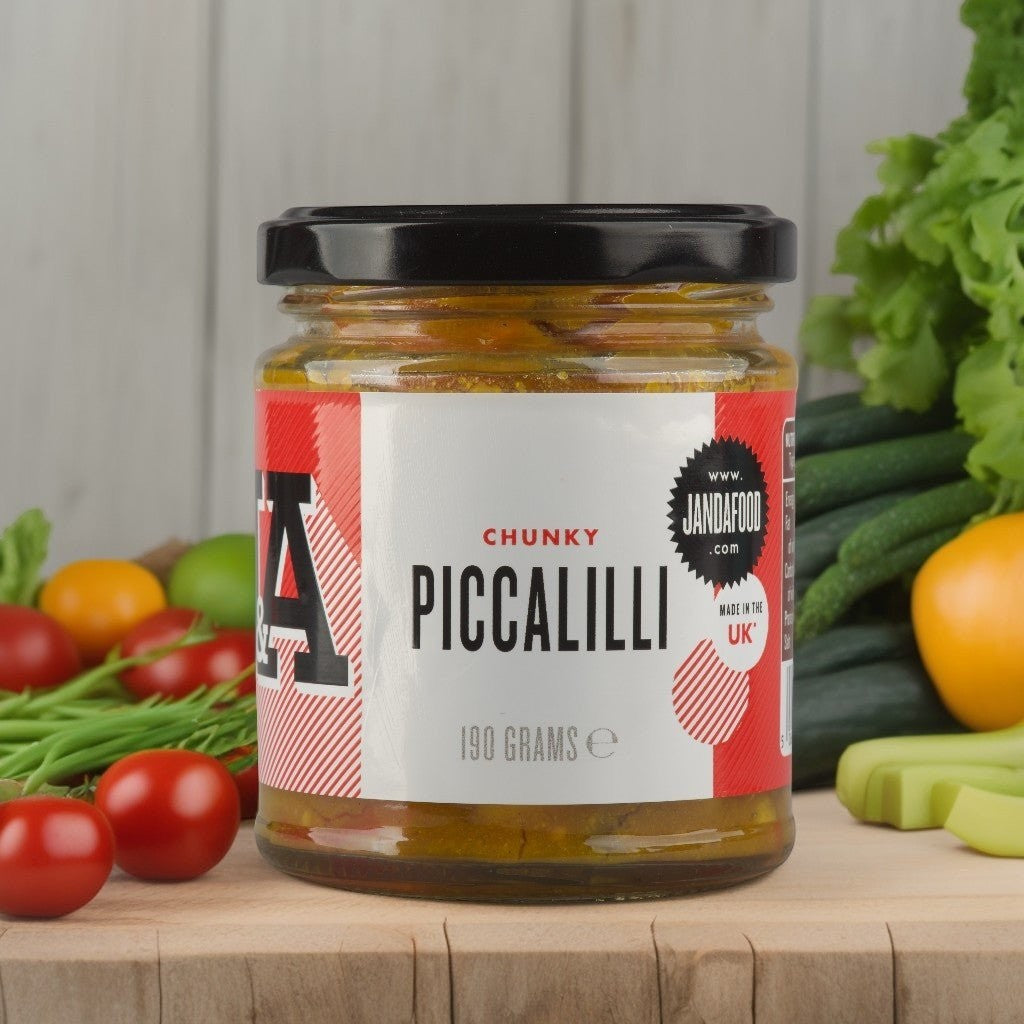 Janda Food Chunky Piccalilli in a glass jar surrounded by fresh vegetables like tomatoes, cucumbers, and peppers.