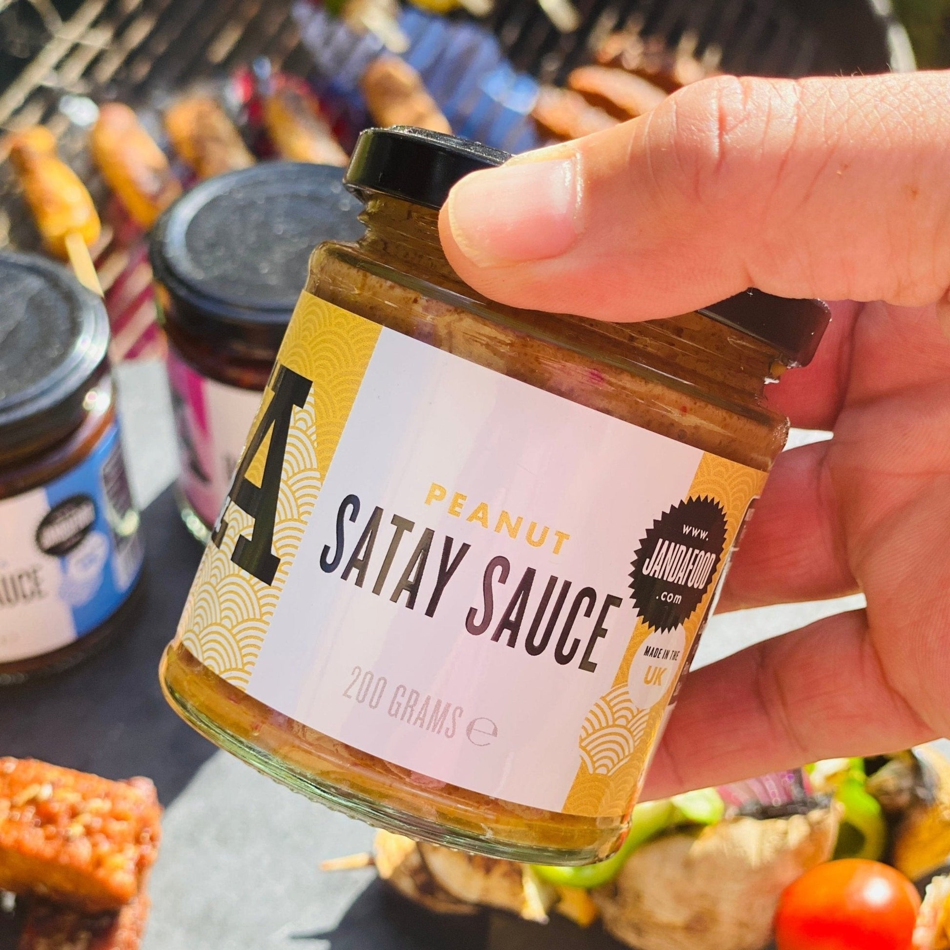 Janda Food Peanut Satay Sauce in a glass jar held by hand over a grill with skewers of meat and vegetables.