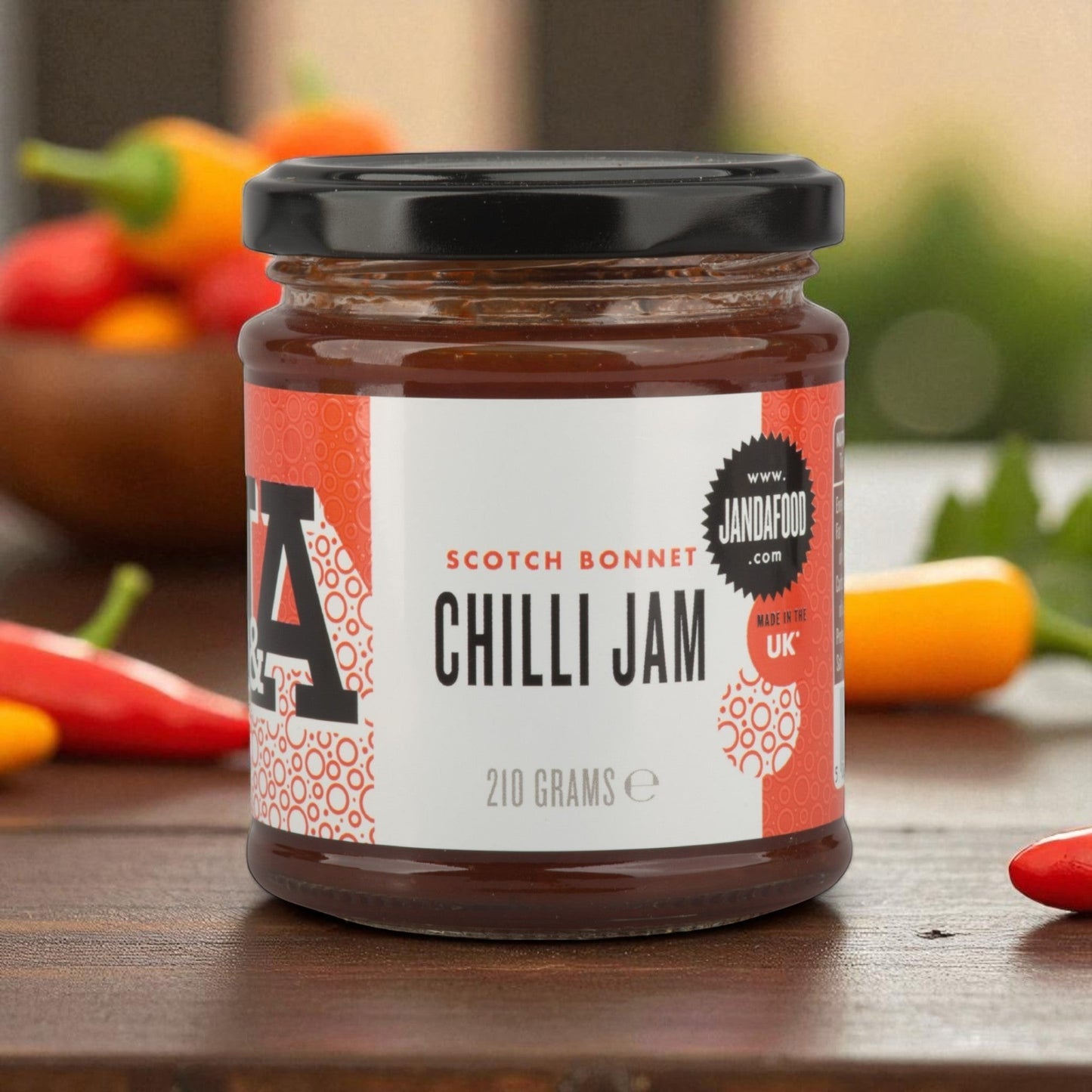 Janda Food Scotch Bonnet Chilli Jam in a glass jar on a wooden table with fresh chillies in the background.