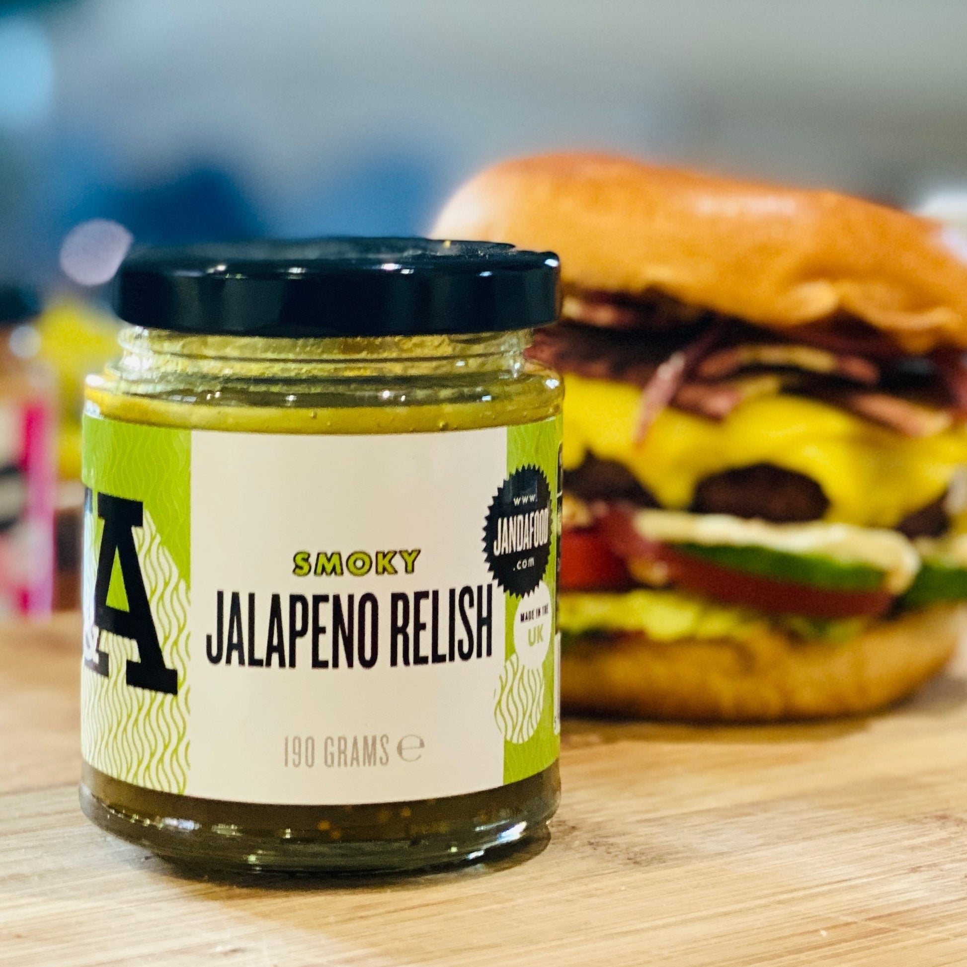Janda Food Smoky Jalapeno Relish jar with a green label, displayed next to a gourmet burger on a wooden board.