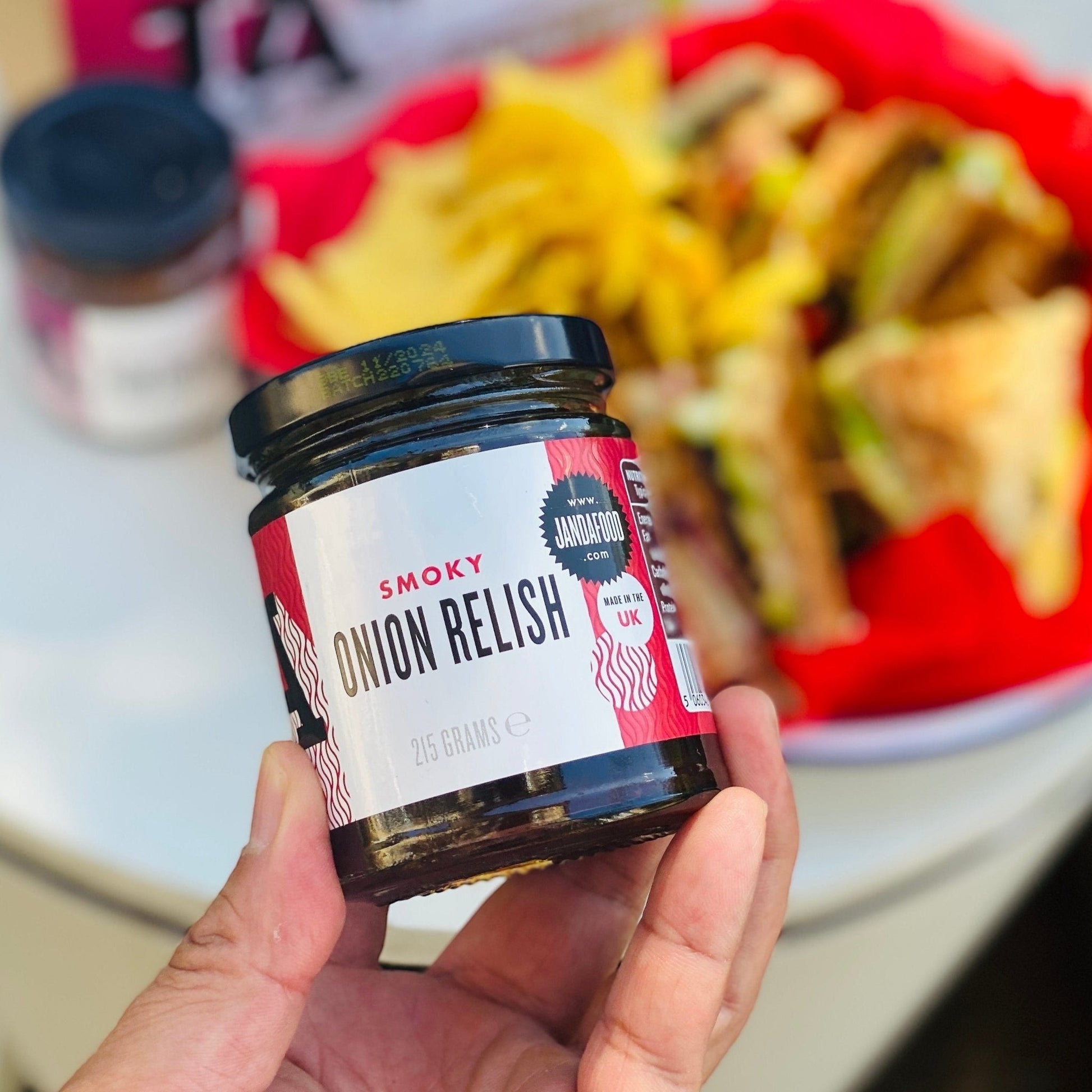 Hand holding a 215g glass jar of Janda Food Smoky Onion Relish with a blurred charcuterie board in the background.