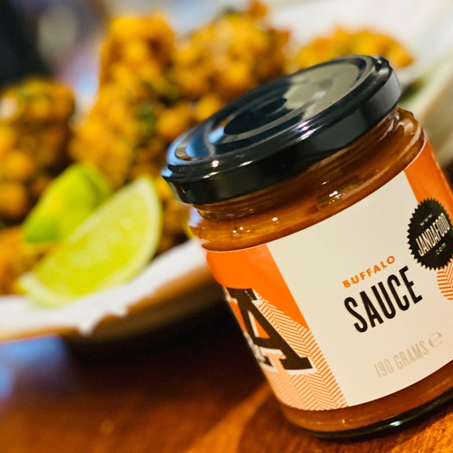 Janda Food vegan buffalo sauce in a glass jar with orange label, served with crispy cauliflower and lime wedges.