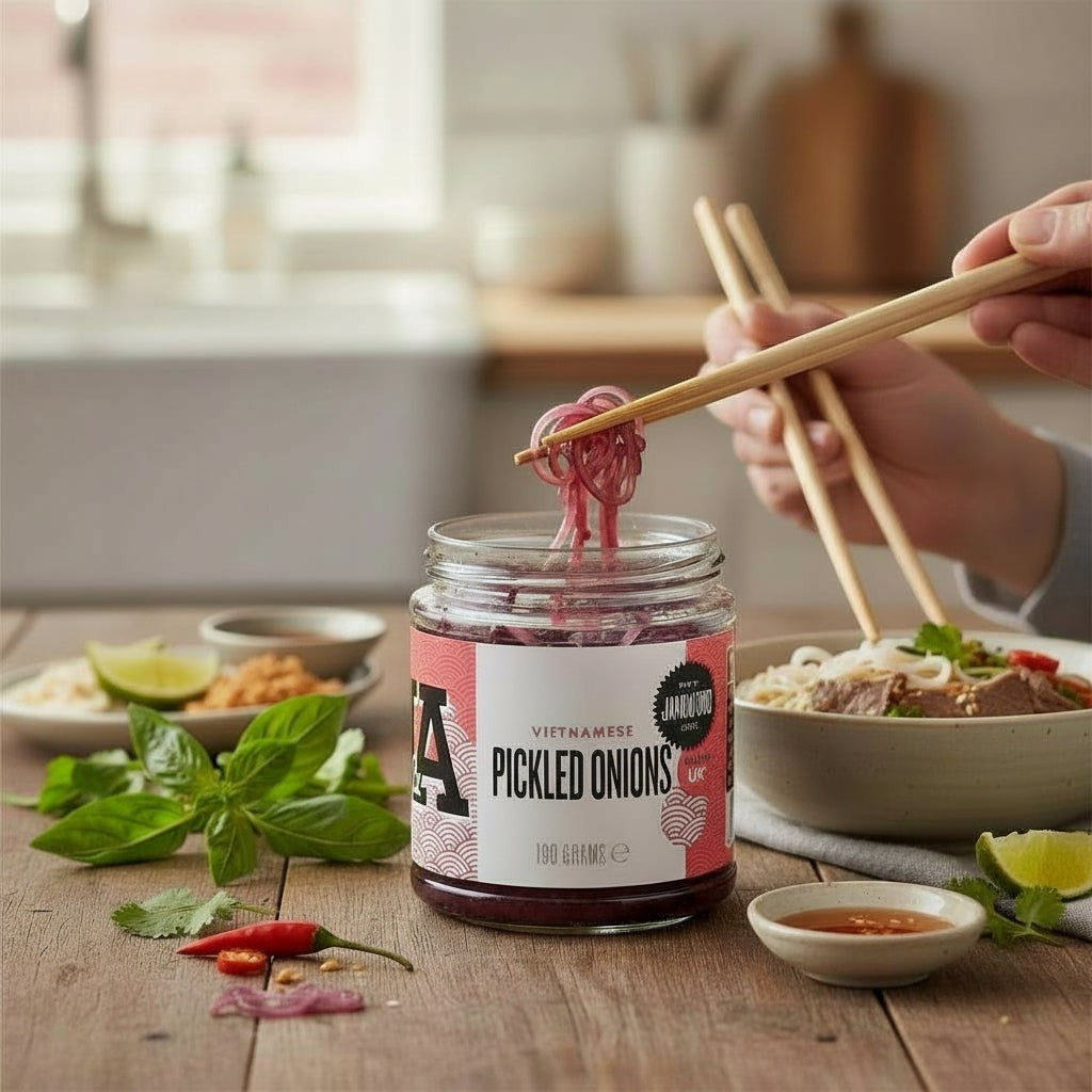 Janda Food Vietnamese pickled red onions in a glass jar with chopsticks lifting a slice over a bowl of pho.